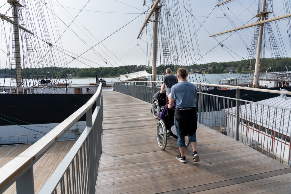 Två unga män syns snett bakifrån, skjutandes på rullstolar med äldre personer i, över Pommerns landgång. De är på väh mot Pommern och är sommarklädda. Det är soligt väder och klar himmel.